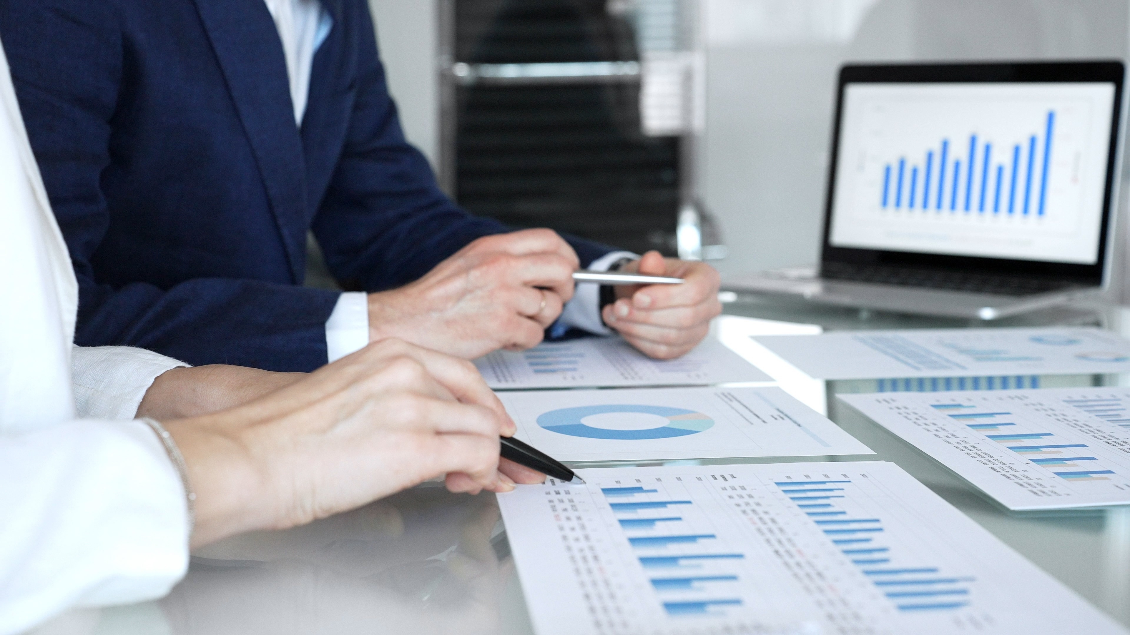 Business professionals collaborating in a modern office, reviewing financial reports, charts and a laptop on a glass table while discussing data analytics, strategy and planning