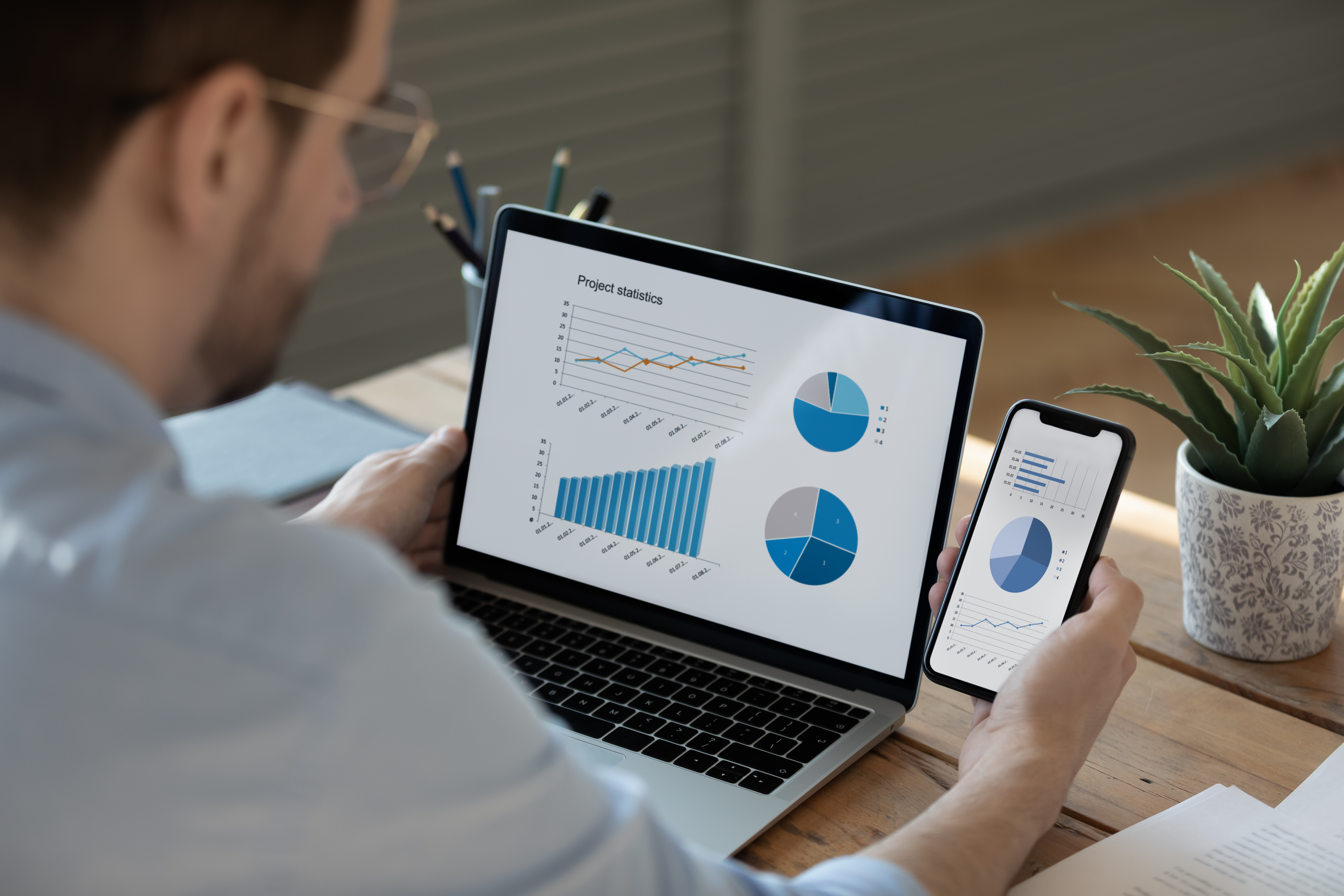 Close up businessman working with statistics, looking at laptop and phone screens with graphs and diagrams, synchronizing electronic devices, checking financial report presentation, sitting at desk